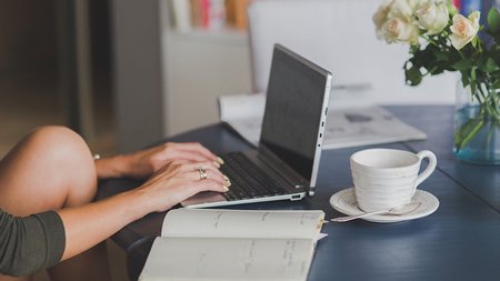 Eine Person sitzt an einem Schreibtisch, vor ihr ein Laptop und eine Tasse.