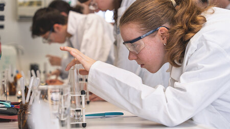 Schülerinnen und Schüler in weißen Kitteln experiomentieren mit Reagenzgläsern im Labor.