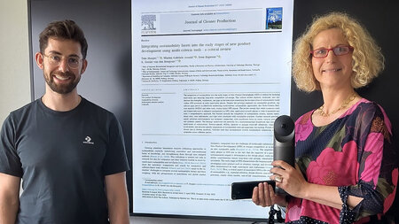 Ein Mann und eine Frau stehen vor einem großen Bildschirm, auf dem eine Publikationsseite aus einer Zeitschrift angezeigt wird.